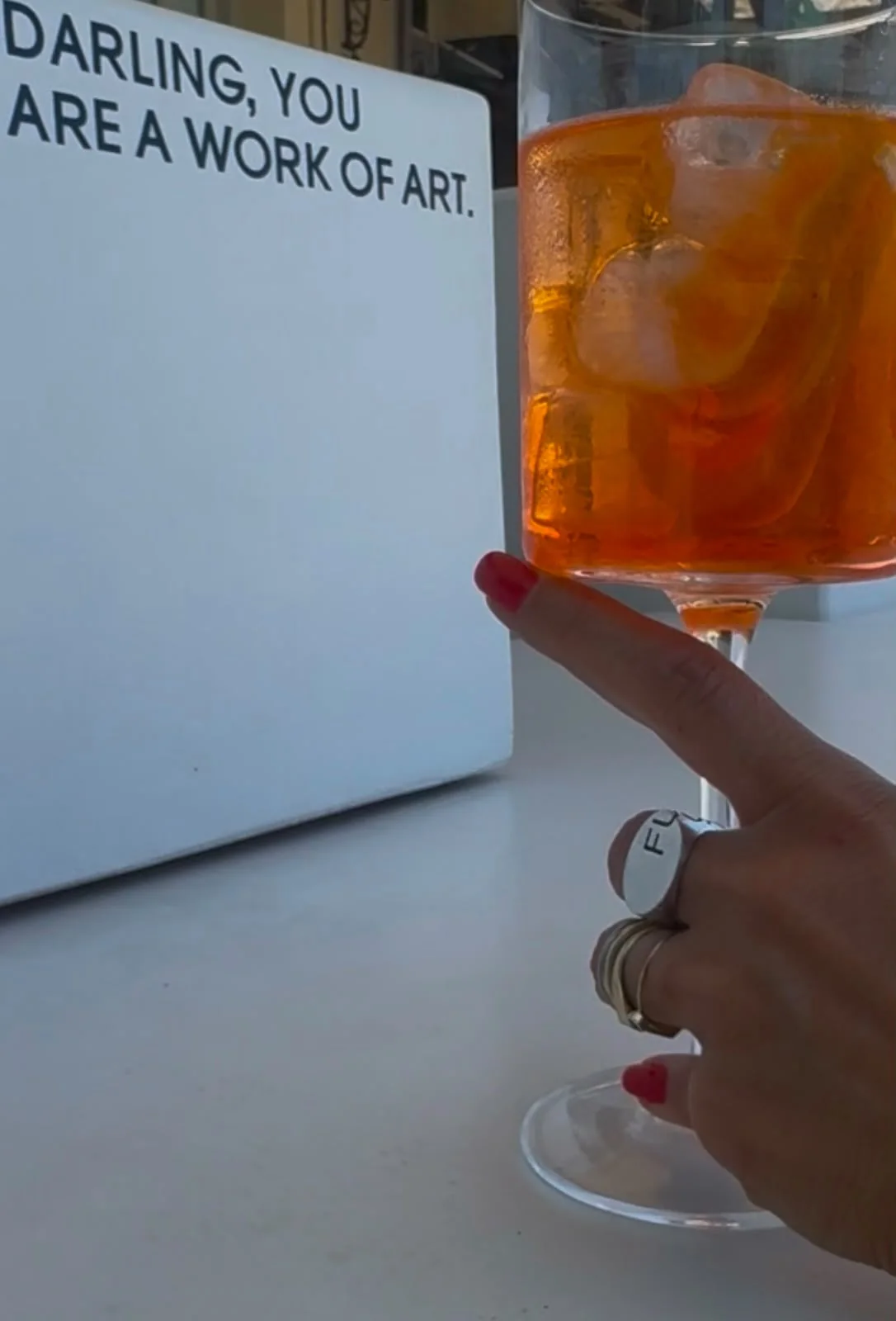 A vibrant orange cocktail beside the Darling You Are A Work of Art sign